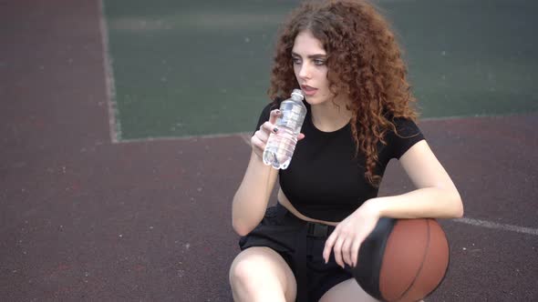 Young Attractive Woman Drinks Water From a Bottle on a Basketball Court During a Break