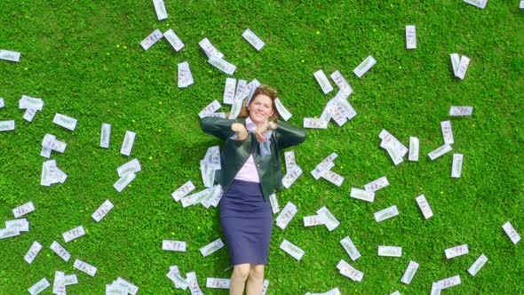 Young Beautiful and Successful Business Lady Lies on a Green Grassy Lawn Strewn with Money