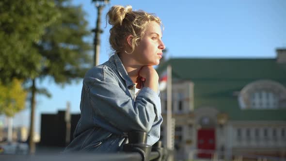 Sideview of Relaxed Attractive Blonde Woman in Denim Jacket Leaning on Fence Blinking As Looking at