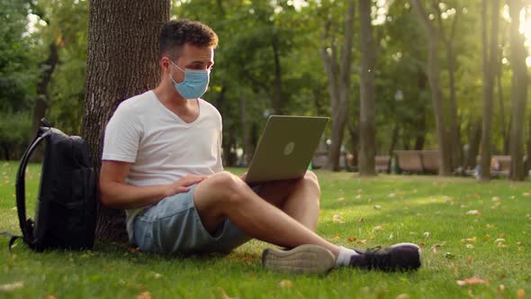 Young Student in a Mask in the Park Sits Under a Tree on the Grass and Communicates Via Video Link