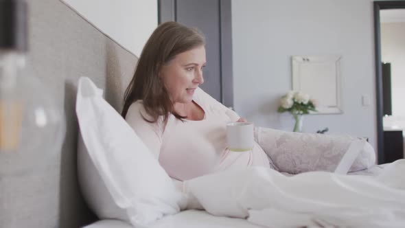 Woman drinking coffee while having a video chat on her laptop in bed at home