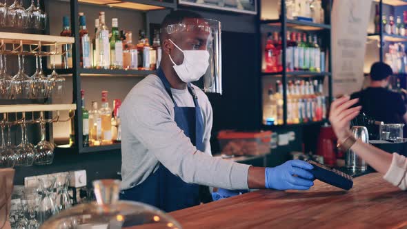 African Ethnicity Waiter in a Face Mask Accepts NFC Payment in the Bar