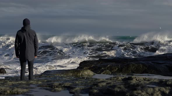 Man Standing On Rocky Shore And Moving To Dodge Crashing Waves