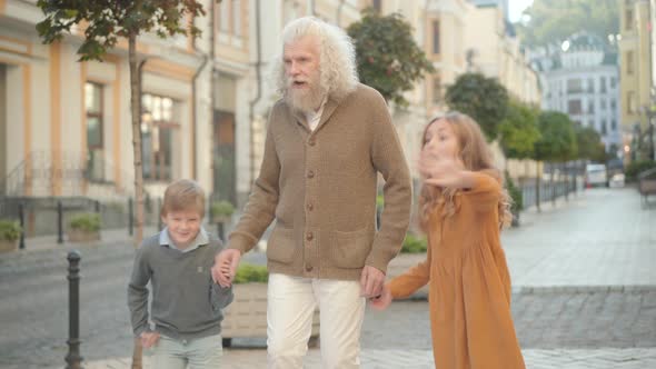 Cheerful Caucasian Brother and Sister Pointing Somewhere and Running Away From Smiling Grandfather