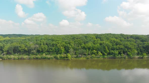 drone flight right of a tree line in thick green forest with blue sky and white clouds on summer sun