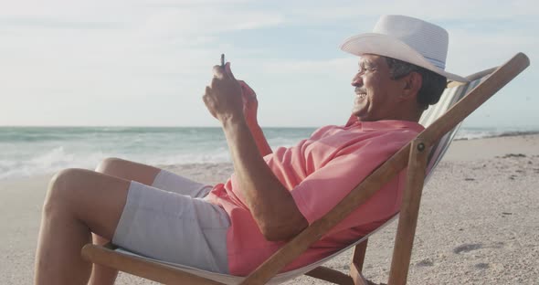 Happy hispanic senior man relaxing on sunbed on beach at sunset, using smartphone