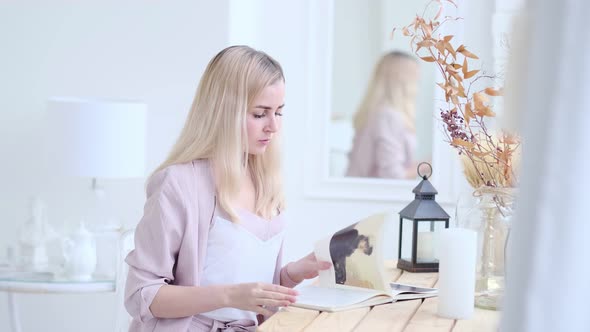 Young Beautiful Woman Reads a Book at the Table in the Living Room