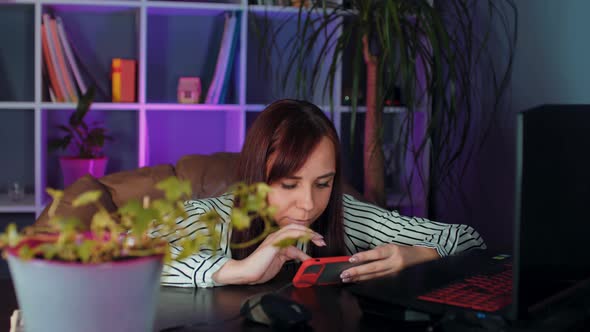 Young Woman Using Smartphone Sitting in Armchair Before Computer in Office