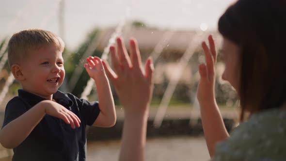 Smiling Boy Waves Hand Splashing Water on Mother in Park