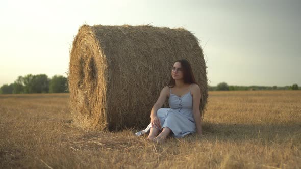 A Young Woman is Sitting Near a Haystack in a Field and a Bee Has Flown to Her