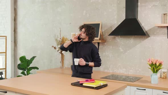 Young Man with Curly Hair is Talking on the Phone While Drinking a Cup of Tea