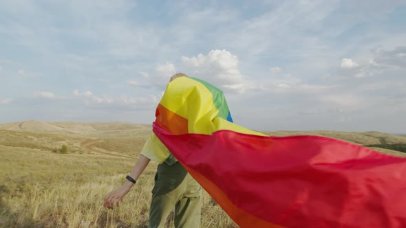 A Thin, Fair-haired Girl with a Developing LGBT Flag on Her Back Walks Across the Field, Against the