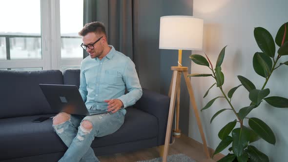 Casual Dressed Man with Glasses Sitting on the Couch and Using Laptop to Make a Video Call From Home