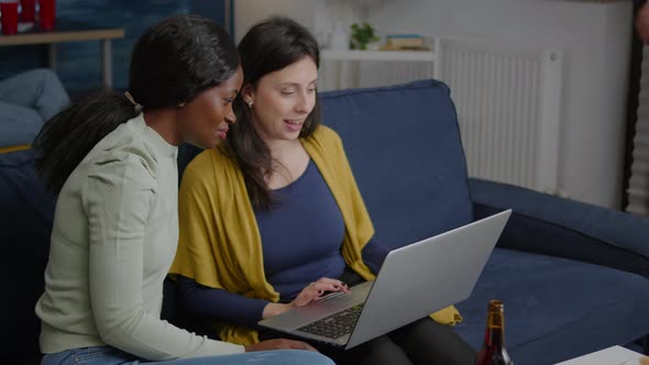 Two Multiethnic Women Sitting on Couch Watching Comedy Series on Laptop