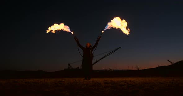 A Man in a Raincoat with Two Flamethrowers Lets Out a Fiery Flame Standing at Sunset on the Sand