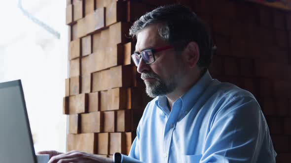 Senior Freelancer Businessman in Glasses Typing Text on the Keyboard in Coffeshop and Drink Coffee