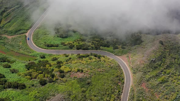 Flying Through the Clouds Over a Mountain Road Surrounded By Green Vegetation. Blue Car Driving on