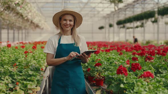Female Gardener Wearing Hat Holding Tablet and Calculate Something Standing Near the Shelf with