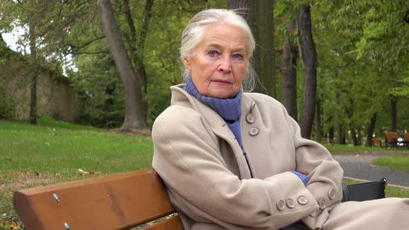 An Elderly Woman Sits on a Bench in a Park and Looks Seriously at the Camera