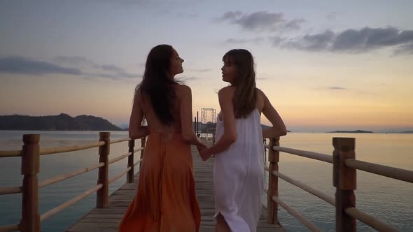 Two Girls in Summer Dresses Running Along the Long Wooden Pier Laughing and Looking Over the