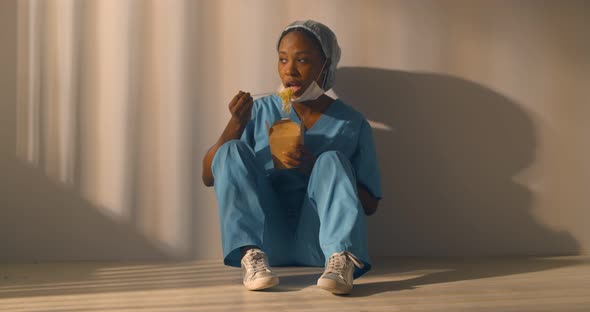 Attractive Afroamerican Nurse in Uniform Sitting on Floor and Eating Takeaway Lunch in Corridor