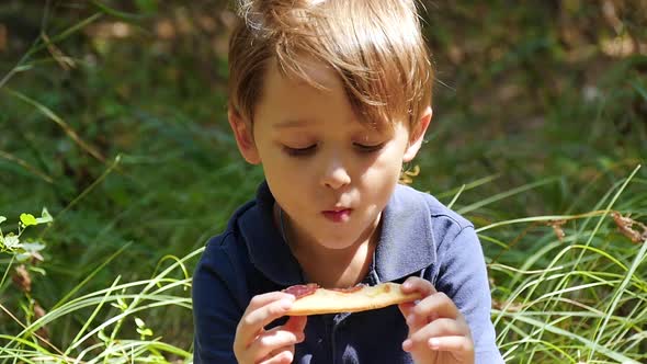 A Boy Bites a Slice of Pizza While Sitting on a Picnic.