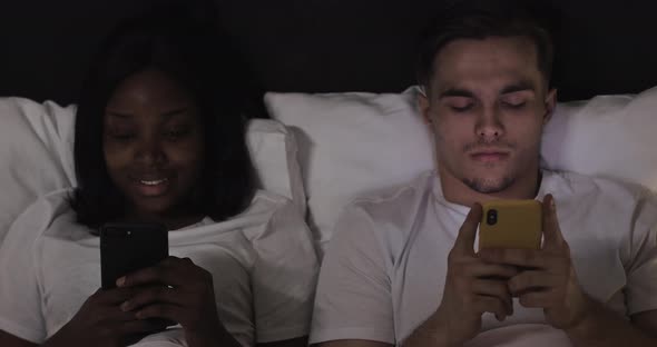 Happy Multi-ethnic Couple with Smartphones in Bed. People, Technology, Internet and Communication