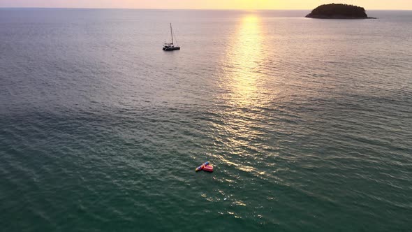 Aerial Flying Over Calm Sunset Waters With Tilt Up View Of Silhouette Of Yacht Floating With Island