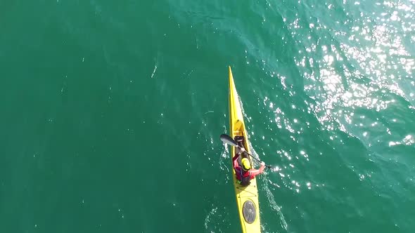 A kayaker paddles in a scenic mountain lake with a drone hovering above him.
