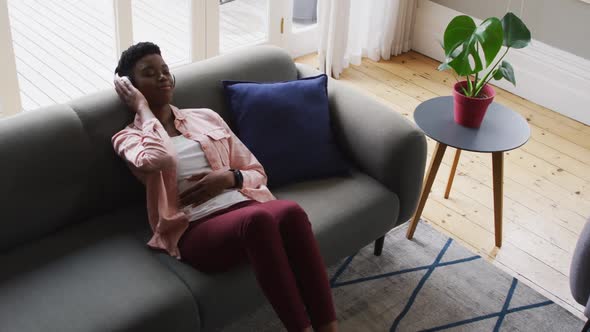 African american woman listening to music on headphones while lying on the couch at home