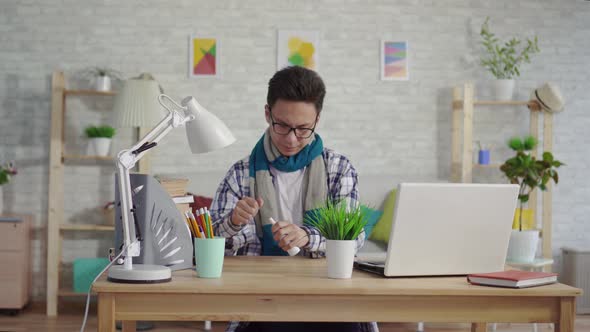 Cold Young Asian Man in a Shirt Uses a Spray for Sore Throat Sitting at the Laptop