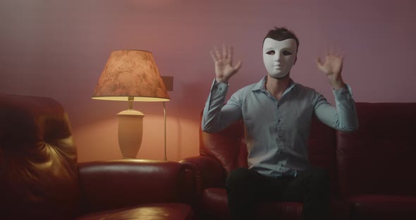 Anonymous man with mask greets with his hand while sitting on the red sofa at home