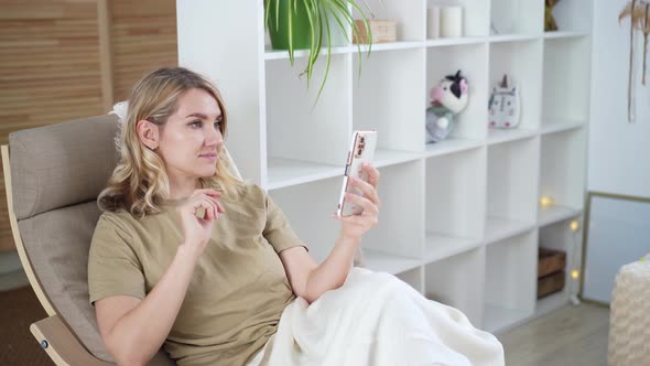 Cute young blonde woman uses phone while sitting at home in armchair Using technology concept
