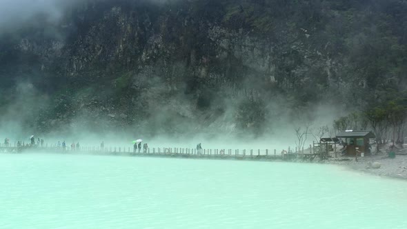 people walking on a bridge with steam rising from sulfur lake and mountain behind, aerial