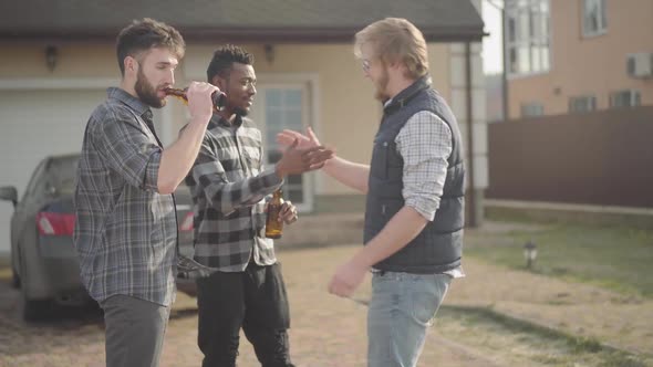 Two Caucasian and African American Men Standing in Front of Big House Drinking Beer. The Third