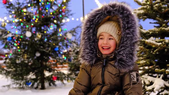 Portrait of happy girl in winter in fur hood against background of fairy lights, Christmas trees