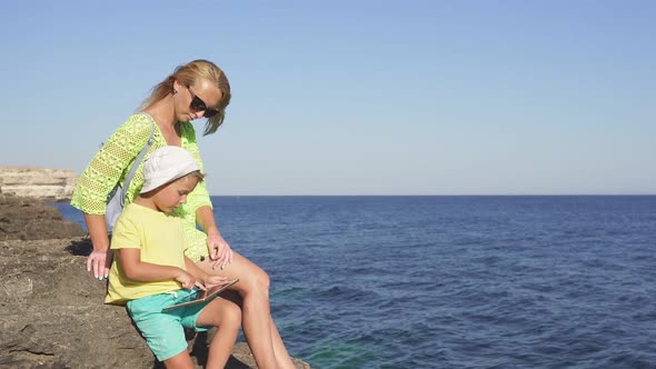 Mother and Child Sit on a Large Rock By the Sea and Use a Tablet