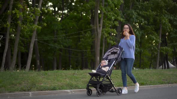 Mom Walks with a Baby in a Stroller and Eats a Burger on the Go