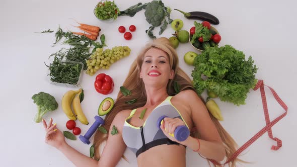 Young Woman Lies Among Fresh Vegetables Holding Dumbbells