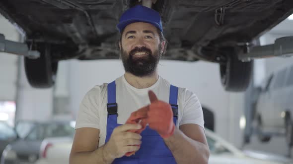 Adult Mechanic Takes Out Gloves Standing Under the Lifted Car and Smiling. Bearded Man in Uniform