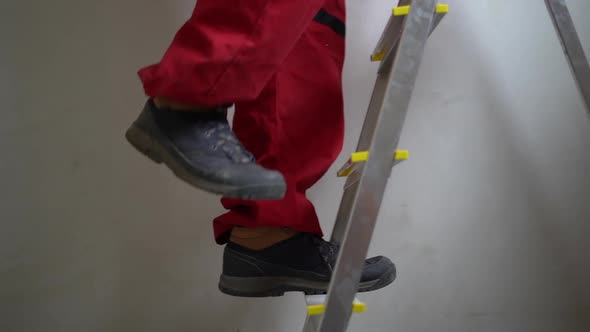 Legs of an Unrecognizable Worker in Red Overalls Climbing a Stepladder