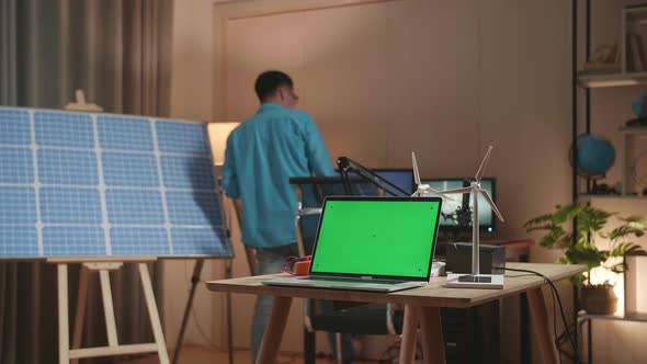 Asian Man Walks Into The Office That Has Solar Cell Next To The Green Screen Laptop And Wind Turbine