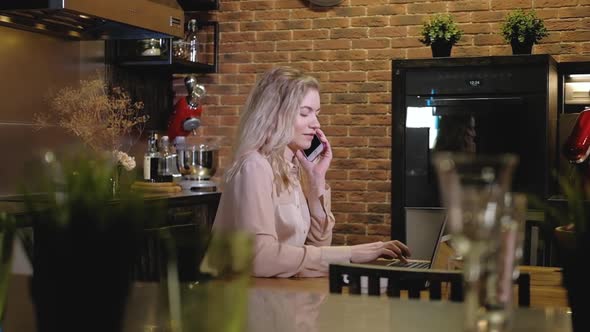 Girl communicates smartphone with client in evening kitchen and type on laptop