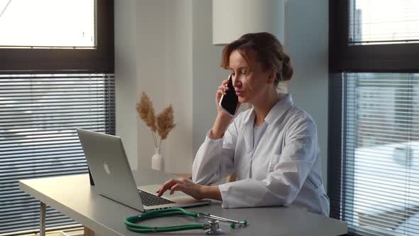 Busy Young Female Doctor in White Coat Typing on Laptop Computer and Talking on Mobile Phone