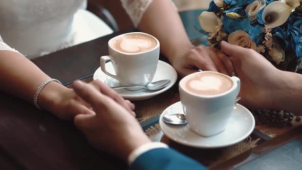 Newlyweds Hold Hands While Sitting in a Cafe, Sitting at a Table on Which There Are Two Mugs of