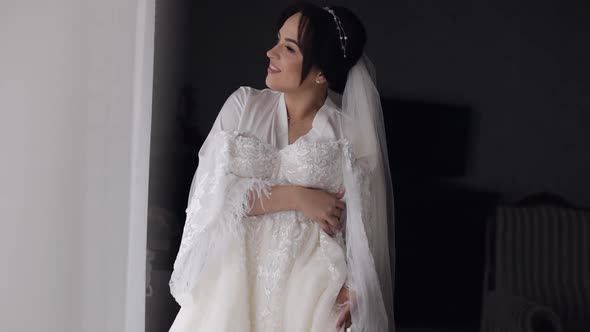 Bride in Lingerie Standing with Her Wedding Dress at Home Near Window White Boudoir Dress and Veil