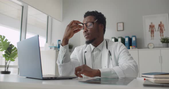 Exhausted African Man Doctor Working on Computer at Desk in Hospital