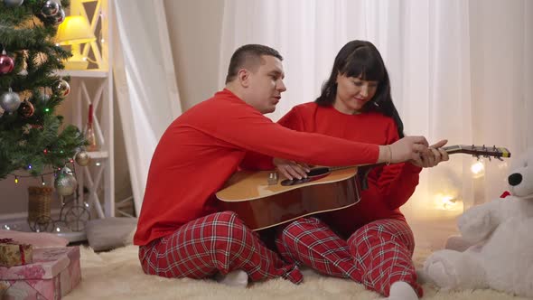 Positive Caring Man Teaching Pregnant Woman Playing Guitar on New Year's Eve at Home