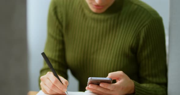 Female executive using mobile phone while writing on diary 4k