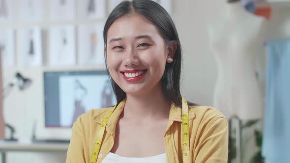 Female Designer With Measuring Tape Around Her Neck Smiling To The Camera While Designing Clothes
